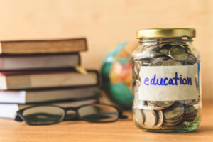 A jar labeled Education with cash inside on a table next to books, glasses, and a globe.