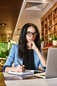 student staring at her computer looking seriously at something as she holds a pen in her hand, writing in a notebook.