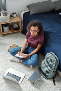 Student taking notes sitting on the floor, leaning up against her bed.