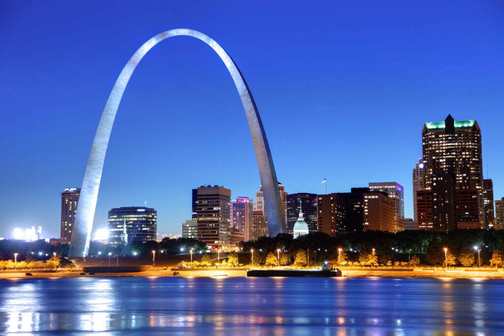 The riverfront of St. Louis showing the Gateway Arch and other buildings at night. There is a slight reflection on the water.