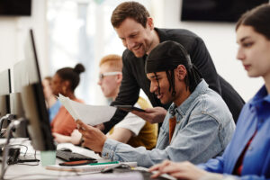 A group of people sitting in front of computers getting help from the person standing up.