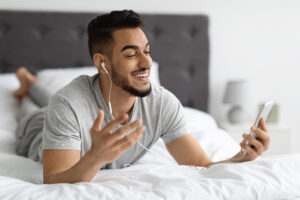 a tan man with a beard lounging in bed with headphones in talking to someone on his phone over facetime.