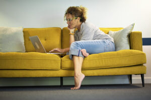 a woman lounging on a yellow couch leaning over to look at her laptop