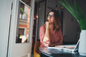 Woman looking out of an open window in front of her computer, pondering the paths she could take in life.