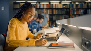 A student in the library working on her computer and taking notes of something.