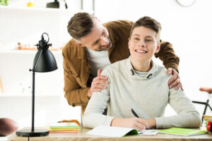 smiling father hugging happy teen son while he doing homework