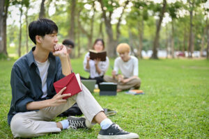 A thoughtful young Asian male college student sits on the grass in the campus park with his book, thinking, pondering, or planning something while looking at the view.