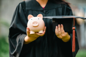 A Graduate holding their graduation hat and a piggy bank.