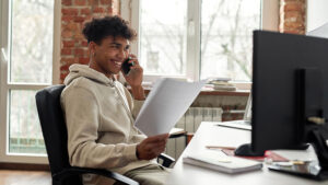 Young student talking on phone, holding a piece of paper and looking at a computer screen. 