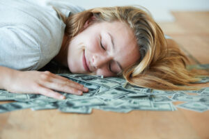 A smiling girl with her eyes closed lying down on the floor with her head in a pile of money.