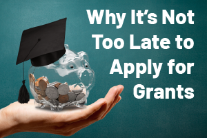 A hand holding a piggy bank that is wearing a graduation hat. The text says "Why it's not too late to apply for grants"